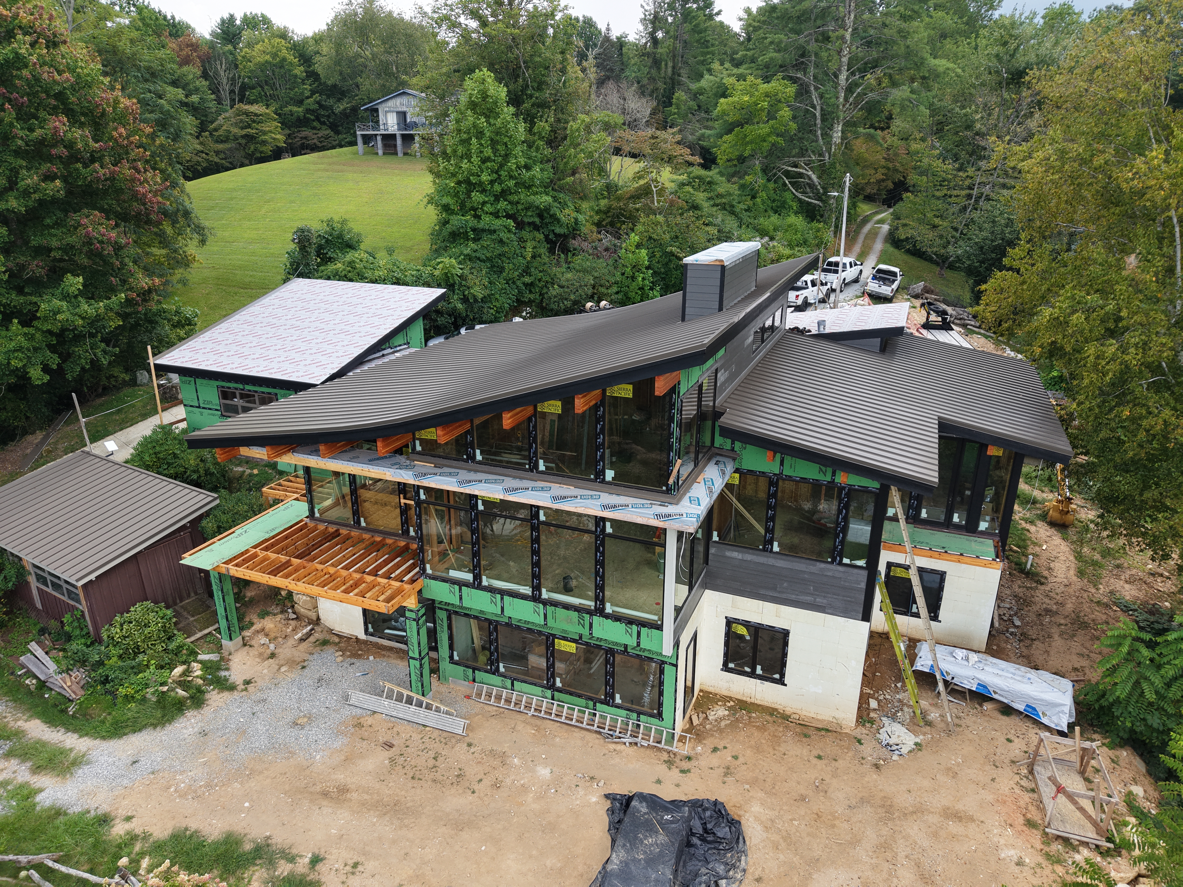 Aerial construction view of the 2026 Executive home with the roofline, glazing, and deck framing in a wooded setting